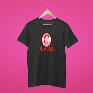 Puede incluir: Camiseta negra con un retrato ovalado rojo y caracteres chinos. El retrato es de una mujer. Los caracteres chinos están en una fuente estilizada. La camiseta está colgada de una percha de madera sobre un fondo rosa.