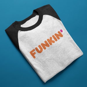 May include: A folded raglan baseball t-shirt with a white body and black sleeves. The shirt displays the word "FUNKIN" in orange letters with a pink dot above the "N". The shirt is on a blue surface.