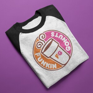 Puede incluir: Camiseta de béisbol raglán blanca y negra con un diseño gráfico circular. El diseño presenta una taza de café y las palabras "Donuts Funkin'" en rosa y naranja. La camiseta está doblada sobre una superficie morada.