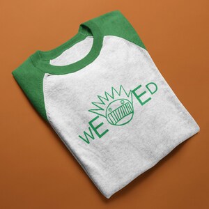 Può includere: Una maglietta da baseball grigia mélange piegata con maniche e colletto verdi. La maglietta presenta una grafica verde di un volto con la scritta "WEED" in verde. La maglietta è esposta su uno sfondo marrone.