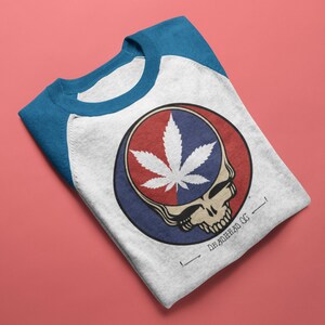 May include: White and blue raglan shirt featuring a graphic of a skull with a cannabis leaf and the text "DEADHEAD OG". The design is in red, white, and blue. The shirt is folded on a pink surface.