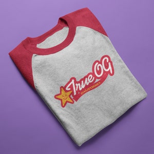 May include: A grey raglan t-shirt with red sleeves and collar. The shirt displays the text "True OG California" in red and white, with a smiling star graphic. The t-shirt is folded on a purple surface.