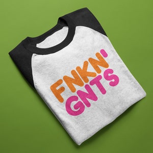 Può includere: Una maglietta da baseball raglan piegata con corpo bianco e maniche nere. La maglietta presenta il testo "FNKN' GNTS" in lettere arancioni e rosa.