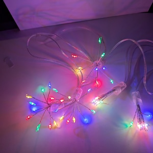 May include: A string of multi-coloured LED fairy lights with red, green, blue, and yellow lights. The lights are arranged in clusters and connected by a clear wire. The lights are on a white surface.