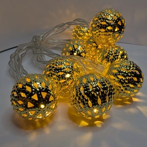 May include: A string of decorative golden globe lights. Each sphere has a textured, perforated design, allowing the warm yellow light to shine through. The lights are strung on a thin, silver wire, creating a festive and elegant look.