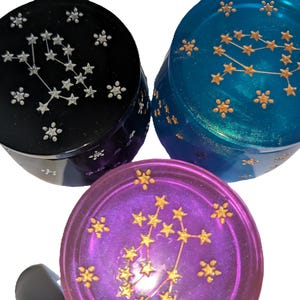 May include: Three round resin trinket boxes with constellation designs. The boxes are black, teal, and purple with gold glitter and gold constellation designs.