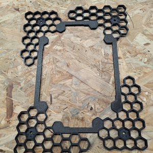 Könnte beinhalten: Ein schwarzer, geometrischer Rahmen mit einem Wabenmuster. Der Rahmen besteht aus einem dunklen Material und hat eine rechteckige Form mit sechseckigen Aussparungen. Das symmetrische und moderne Design eignet sich für Heimdekoration oder Bastelprojekte.