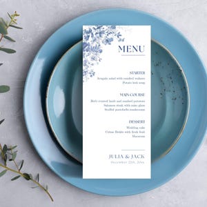 May include: A wedding menu card with a blue floral design, placed on a light blue plate. The menu includes starters, main courses, and desserts. The names "Julia & Jack" and the date "December 22th, 20xx" are also printed on the card.