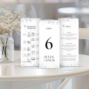 May include: A wedding table number card with a white background and a leafy green design. The card features the number "6" in black text, the names "Julia and Jack" in black text, and a wedding timeline with icons and times.