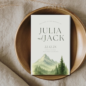 May include: Wedding invitation with a watercolor illustration of a mountain range and pine trees. The text reads "Welcome to the wedding of Julia and Jack 22.12.2X Little Chapel Boulder Colorado."