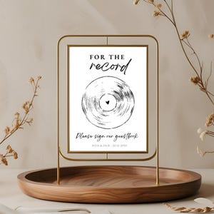 Puede incluir: Un letrero con marco de metal dorado con el texto "FOR THE record" sobre una ilustración de disco de vinilo en blanco y negro. Debajo de la ilustración está el texto "Please sign our guestbook". El letrero está sobre una bandeja de madera.