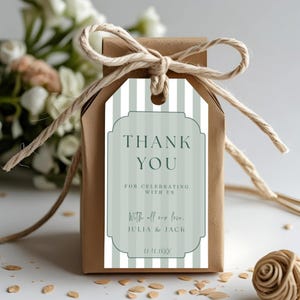 May include: A small, brown gift box tied with twine, featuring a tag with the words "THANK YOU." The tag has a striped background in shades of green and white, with the names "JULIA & JACK" and a date at the bottom. The box is decorated with a twine bow.