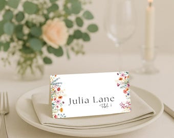 Peut inclure: Une carte de place blanche avec des motifs floraux et le nom "Julia Lane" imprimé en noir. La carte est posée sur une assiette blanche avec une serviette pliée, avec un arrangement floral et une bougie allumée en arrière-plan.