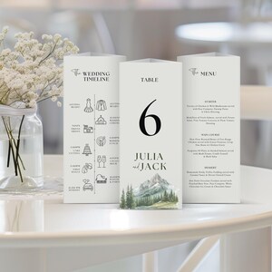Op de afbeelding: Drie witte trouwtafelkaarten. Eén kaart toont een bruilofttijdlijn met illustraties. Een andere kaart toont het nummer 6, de namen "Julia en Jack" en een berglandschap. De derde kaart bevat een menu met voorgerechten, hoofdgerechten en desserts.