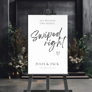 May include: A white wedding welcome sign on a wooden easel. The sign reads "All because two people swiped right" with the names Julia & Jack and the wedding date. The sign is surrounded by floral arrangements.