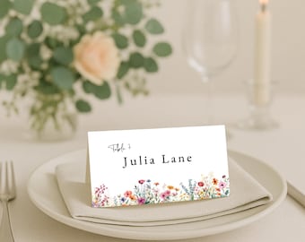 Peut inclure: Une carte de place blanche avec le texte "Table 7" et "Julia Lane" en noir. Le bord inférieur présente un motif floral à l'aquarelle dans les tons de rose, d'orange, de violet et de jaune. La carte repose sur une assiette blanche avec une serviette pliée.