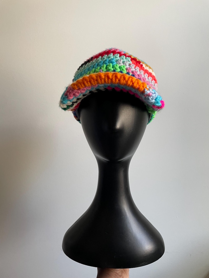 May include: A multi-colored crochet bucket hat featuring a vibrant mix of yarn colors including bright orange, pink, blue, green, and white. The hat has a textured, hand-stitched appearance with a wide, slightly downturned brim and a rounded crown.