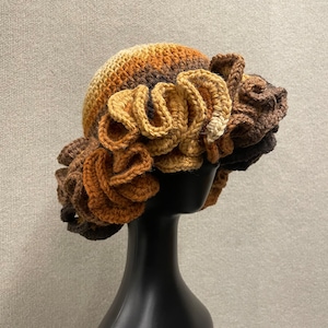 May include: A handmade crochet hat in shades of brown, tan, and black, featuring a ruffled brim. The hat is displayed on a black mannequin head, showcasing its unique texture and design. This hat is a stylish accessory.