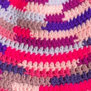 May include: A multi-colored crochet fabric featuring a dense, textured pattern of interlocking yarn loops. The design displays a vibrant mix of hot pink, deep purple, light lavender, soft peach, and muted taupe colors arranged in horizontal rows.