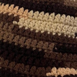 May include: A close-up view of a crocheted textile featuring horizontal stripes in varying shades of brown, dark brown, and beige. The fabric displays a consistent, repeating stitch pattern throughout, creating a textured surface with a dense, woven appearance.