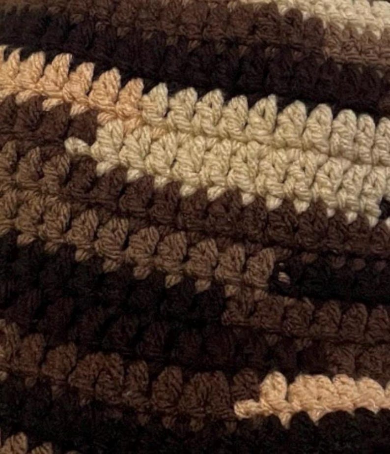 May include: A close-up view of a crocheted textile featuring horizontal stripes in varying shades of brown, dark brown, and beige. The fabric displays a consistent, repeating stitch pattern throughout, creating a textured surface with a dense, woven appearance.
