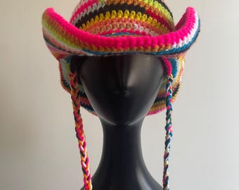 May include: A multi-colored crochet cowboy hat featuring horizontal stripes in neon pink, lime green, orange, black, white, and blue. The hat has a curved brim and two long, braided yarn chin straps that hang down from the sides of the crown.