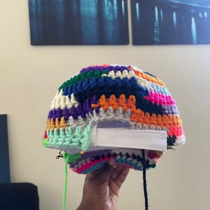 May include: A baseball cap featuring a multi-colored crocheted yarn overlay in shades of neon pink, orange, yellow, green, blue, purple, and cream. The cap includes a white fabric adjustable strap at the back with loose yarn ends hanging down.