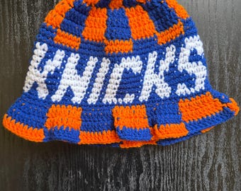 May include: A crochet bucket hat featuring a checkered pattern in vibrant orange and royal blue. The word KNICKS is stitched in white yarn across the center of the hat. The design uses a thick yarn texture with a rounded crown and a wide brim.