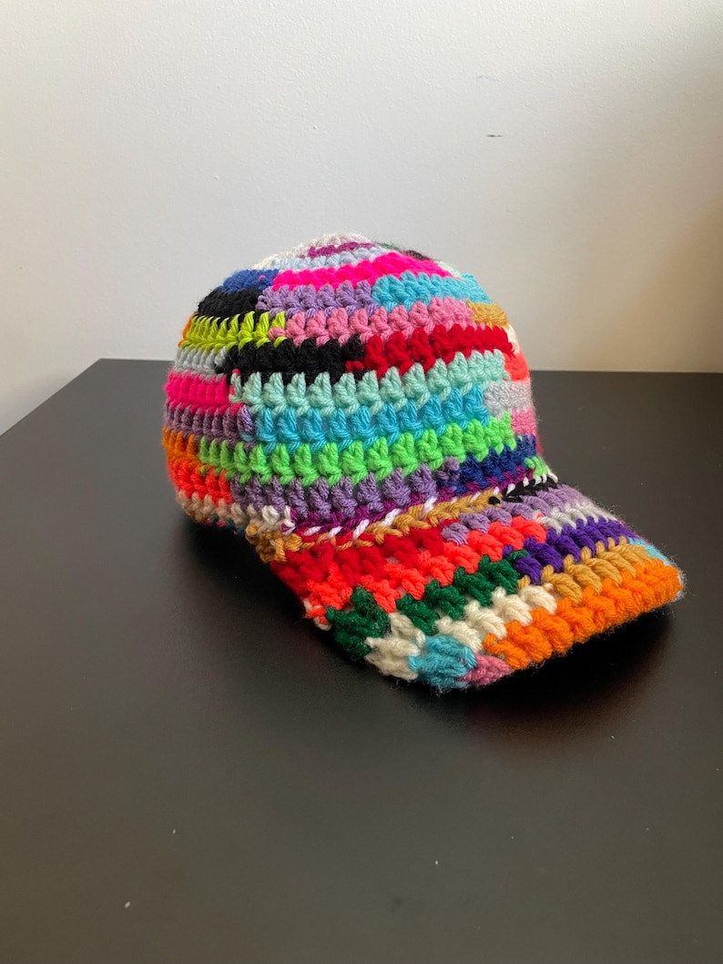 May include: A baseball cap crafted from multicolored crochet yarn featuring a vibrant patchwork design. The hat displays a variety of bright hues including neon pink, electric blue, lime green, orange, purple, black, and white in a textured stitch.
