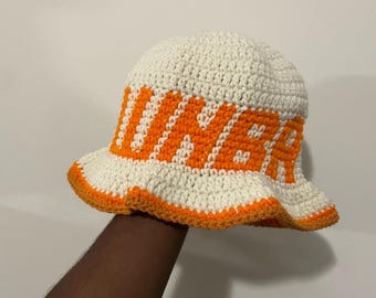 May include: A cream-colored crochet bucket hat featuring the word "UNER" in bold orange lettering around the crown. The hat has a rounded top and a wide, slightly flared brim with a vibrant orange trim along the bottom edge of the brim.