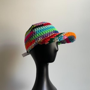 May include: A baseball cap made of multi-colored crocheted yarn featuring horizontal stripes in vibrant shades of red, orange, yellow, green, blue, purple, and black. The cap has a structured crown and a curved brim, with a small white tag visible.