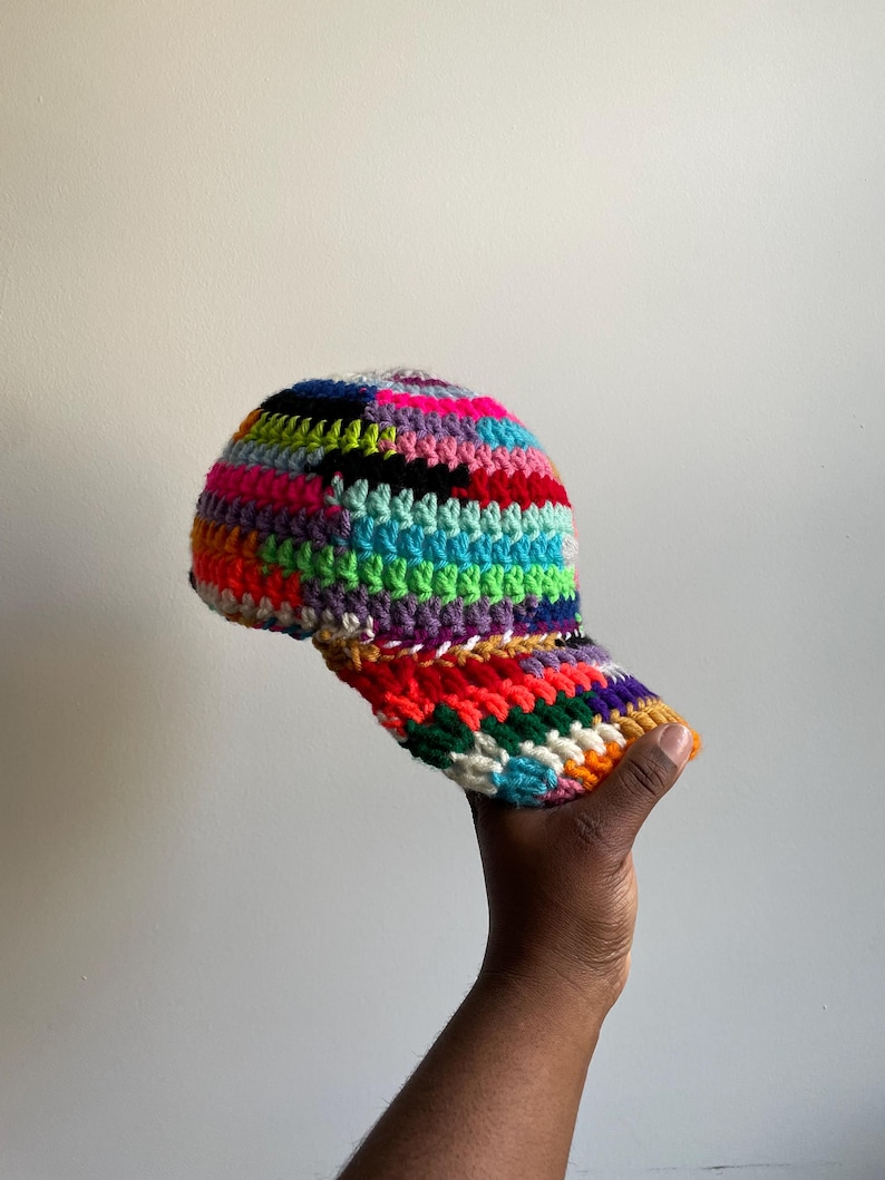 May include: A baseball cap constructed from multicolored crocheted yarn featuring a vibrant palette of neon pink, bright blue, lime green, purple, orange, and black. The cap has a structured crown and a curved brim, all worked in a textured stitch.