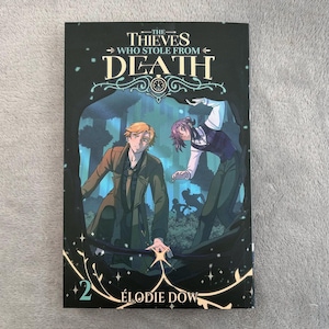 Può includere: Copertina del libro "The Thieves Who Stole From Death" di Élodie Dow. La copertina presenta due figure maschili in un ambiente forestale. Il titolo è in bianco e oro, con il nome dell'autore e il numero "2" in basso.
