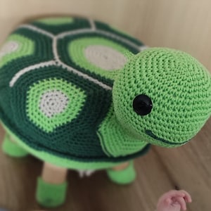 May include: A green crocheted turtle stool with a white and green shell pattern. The turtle has black eyes and a smiling mouth.