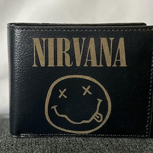 Op de afbeelding: Zwart bi-fold portemonnee met het woord "NIRVANA" in gouden letters. Onder het woord staat een gouden smiley met doorgestreepte ogen en een uitgestoken tong. De portemonnee is gemaakt van een textuurrijk materiaal.