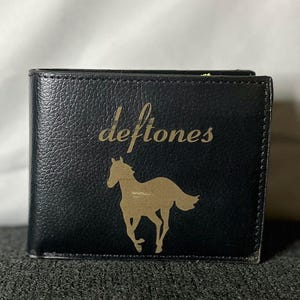 May include: Black bi-fold wallet with the band name "deftones" in gold lettering and a gold horse silhouette. The wallet is made of a textured material.