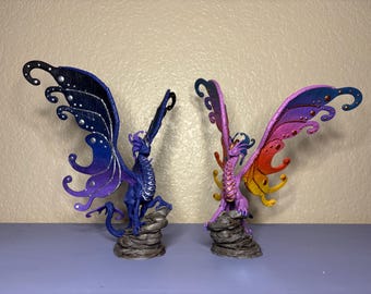 May include: Two painted dragon figurines with butterfly wings. The dragon on the left has blue, purple, and silver wings. The dragon on the right has pink, orange, and yellow wings.