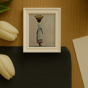 May include: A framed painting depicts a person carrying a basket of fruit on their head. The artwork is in a white frame with a beaded border. The person is wearing a white dress and apron. The painting is set against a neutral background.