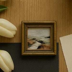 May include: Small framed landscape painting with a gold-colored frame. The artwork depicts a river scene with muted tones of green, brown, and white. The painting is displayed on a wooden surface, with white tulips and a dark gray cloth nearby.