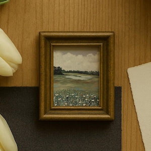May include: A small framed landscape painting with a gold-colored frame. The artwork depicts a serene scene of a river, green fields, and a cloudy sky. White flowers dot the foreground. The painting is displayed on a wooden surface.
