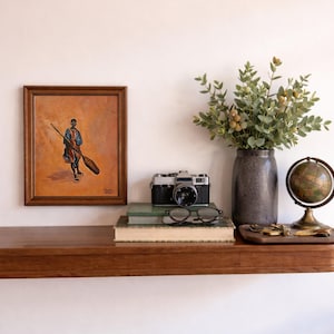 May include: A wooden shelf displays a framed painting, a vintage camera, books, glasses, a vase with greenery, and a globe. The painting depicts a figure holding a broom. The camera is black and silver.
