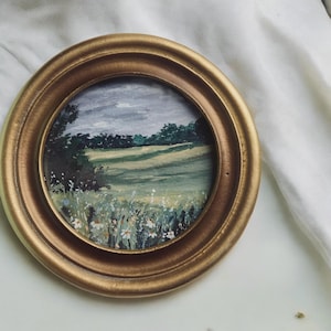May include: A small, round, gold-framed painting of a landscape. The artwork depicts a field with green and yellow hues, wildflowers, and a tree line under a cloudy sky. The frame has a simple, classic design.
