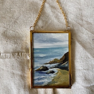 May include: A rectangular painting of a coastal landscape featuring a sandy beach, dark rocky cliffs, and blue ocean waves under a cloudy sky, encased in a thin gold metal frame with a matching gold chain for hanging.