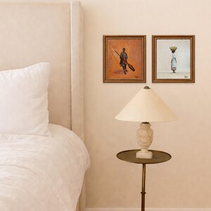 May include: Two framed paintings depict figures with a warm, earthy palette. One shows a person with a broom, the other a person carrying a basket. A cream-colored lamp with a round, brass-toned table sits below the artwork.