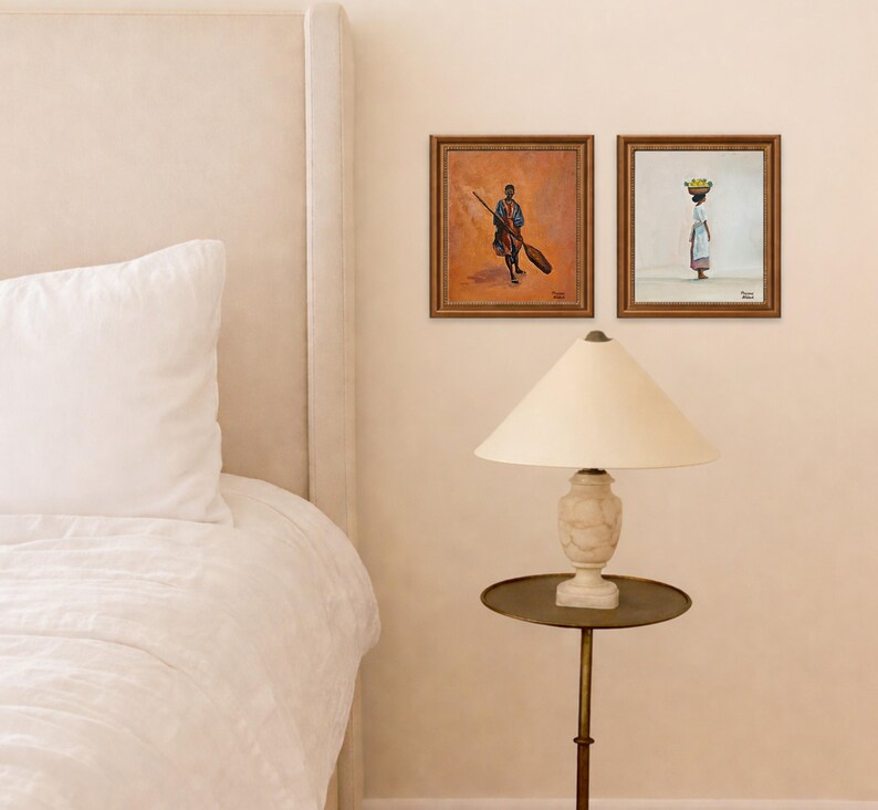 May include: Two framed paintings depict figures with a warm, earthy palette. One shows a person with a broom, the other a person carrying a basket. A cream-colored lamp with a round, brass-toned table sits below the artwork.