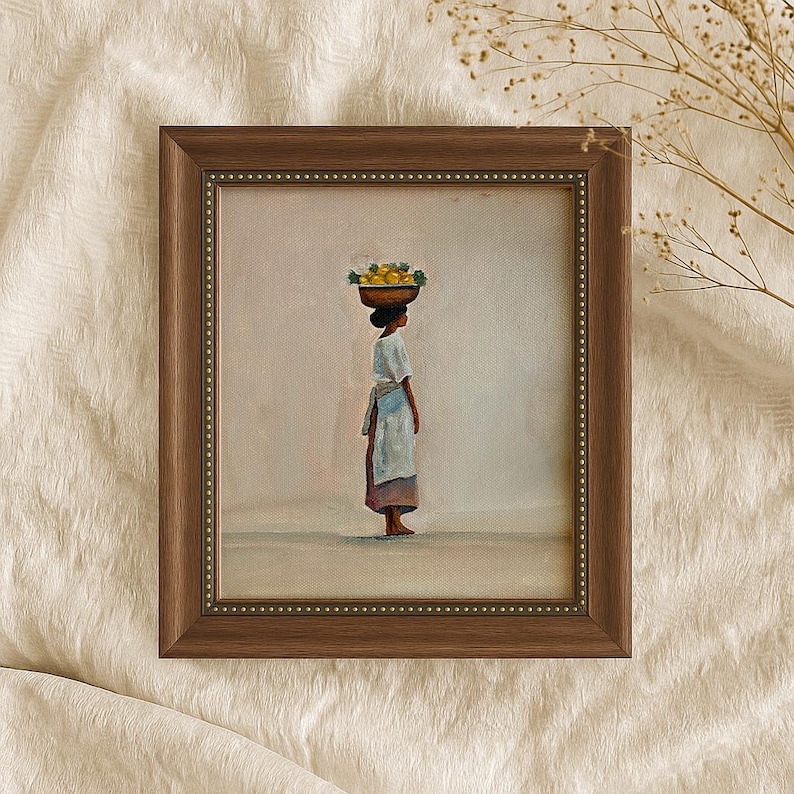 May include: Framed painting of a person carrying a basket of yellow fruit on their head. The person is wearing a white dress with a blue apron. The painting is in a brown frame with a beaded border.