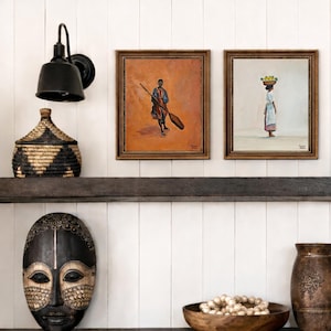 May include: Two framed paintings depicting figures in traditional attire. One shows a person with a paddle, the other a person carrying a basket on their head. A decorative mask, a woven basket, and a wooden bowl with beads are also visible.