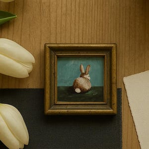 May include: A small framed painting of a brown rabbit with a white tail, set against a teal background. The painting is in a gold-coloured frame. The image is on a wooden surface, with white tulips and a dark grey cloth.