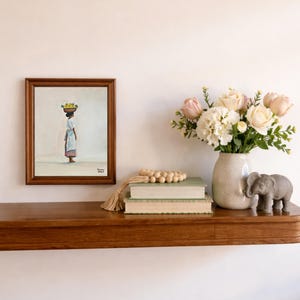 May include: A framed painting depicts a person carrying a basket of fruit on their head. A vase of flowers, two books, a wooden bead garland, and a small elephant statue are on a wooden shelf. The shelf is mounted on a white wall.