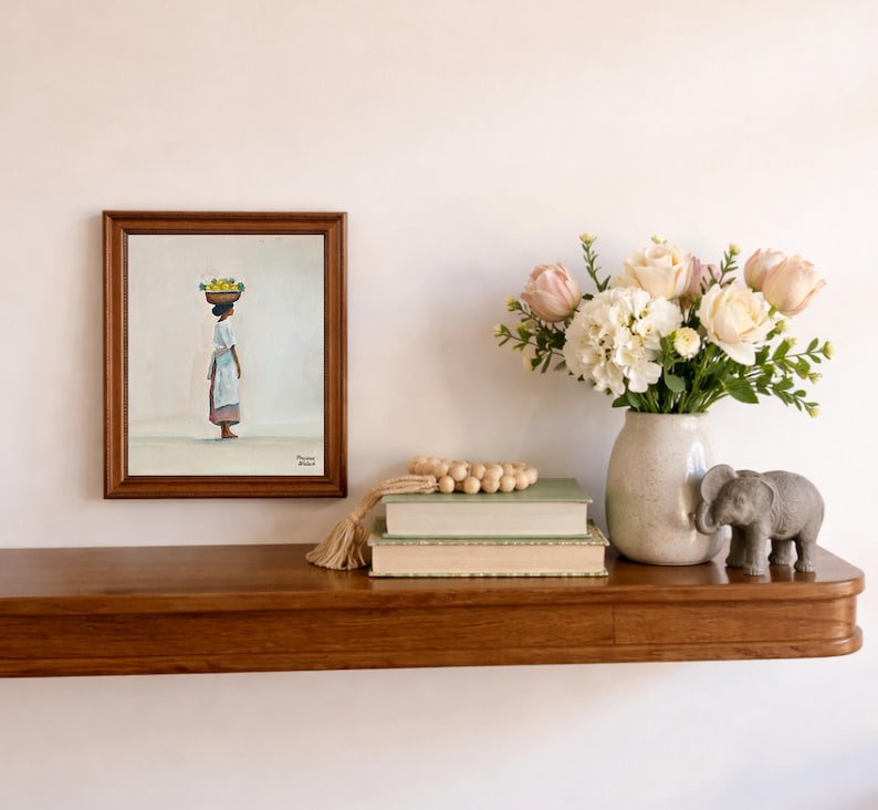 May include: A framed painting depicts a person carrying a basket of fruit on their head. A vase of flowers, two books, a wooden bead garland, and a small elephant statue are on a wooden shelf. The shelf is mounted on a white wall.